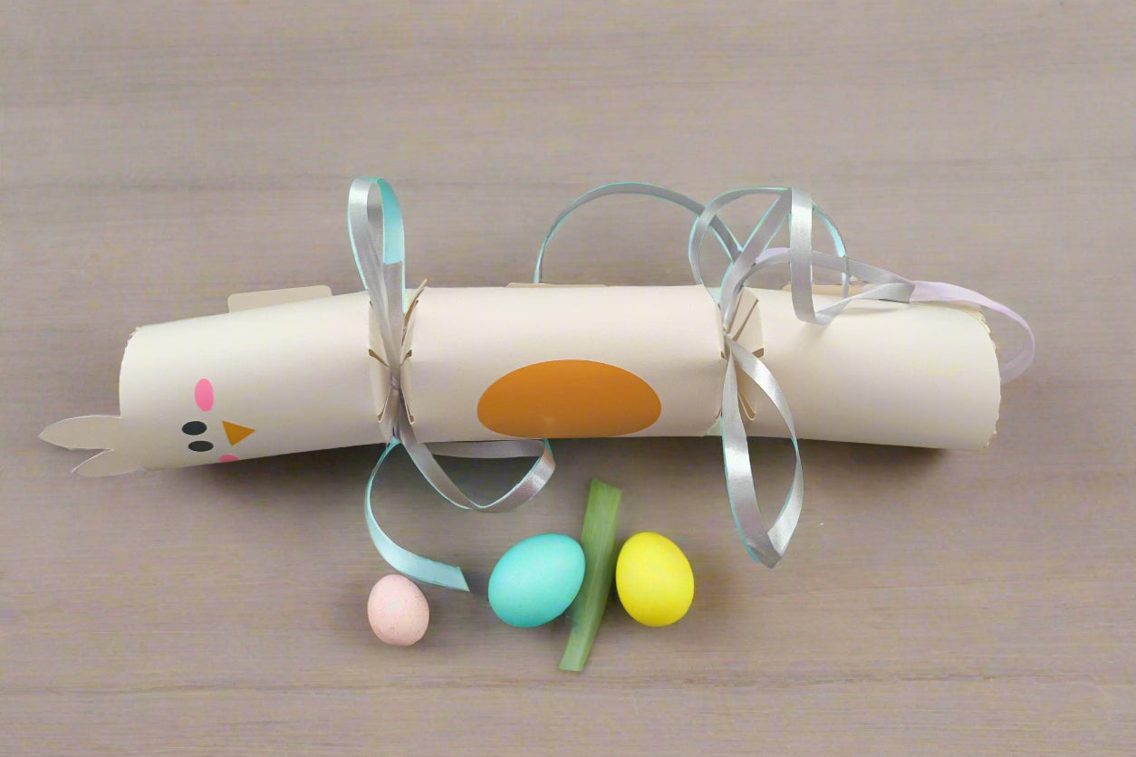 Cream Easter chick cracker with orange oval belly and silver ribbon ties, displayed flat on a table beside pastel mini eggs for size reference.