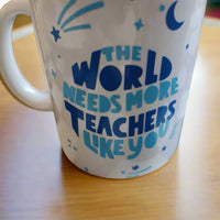 A white ceramic mug with a handle on the left side, featuring blue text that reads, "THE WORLD NEEDS MORE TEACHERS LIKE YOU." The design is complemented by small blue decorative elements, including stars, a crescent moon, and abstract shapes. The mug rests on a wooden surface, creating a simple and heartfelt presentation, perfect for celebrating teachers' contributions.