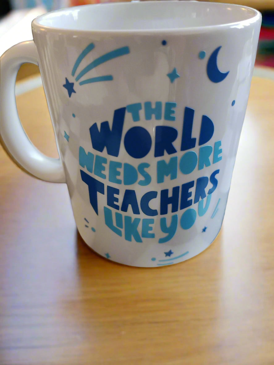 A white ceramic mug with a handle on the left side, featuring blue text that reads, "THE WORLD NEEDS MORE TEACHERS LIKE YOU." The design is complemented by small blue decorative elements, including stars, a crescent moon, and abstract shapes. The mug rests on a wooden surface, creating a simple and heartfelt presentation, perfect for celebrating teachers' contributions.