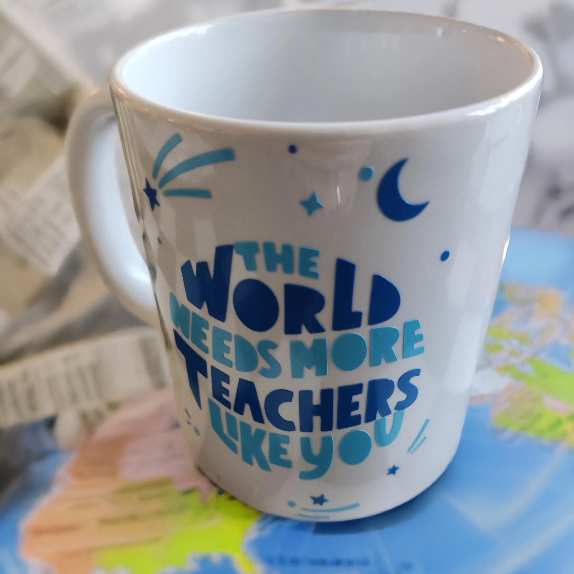 A white ceramic mug with blue text that reads, "THE WORLD NEEDS MORE TEACHERS LIKE YOU." The design is adorned with simple blue decorative elements, including stars and abstract shapes. The mug is placed on a colourful world map, symbolising the global importance of teachers and education, with a blurred background adding a subtle yet warm effect. Perfect for celebrating and appreciating educators.