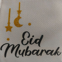 Close-up of a white textured hessian fabric with gold glitter moon and star decorations and the black text 'Eid Mubarak' in elegant script.