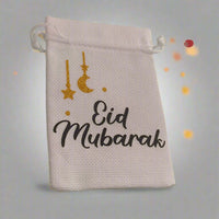White hessian drawstring bag with 'Eid Mubarak' text in black and gold glitter crescent moon and stars, placed against a soft grey background with glowing orange bokeh lights.