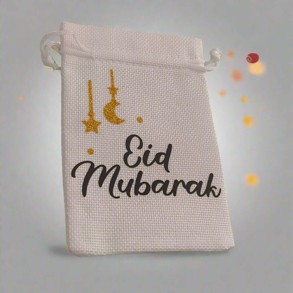 White hessian drawstring bag with 'Eid Mubarak' text in black and gold glitter crescent moon and stars, placed against a soft grey background with glowing orange bokeh lights.