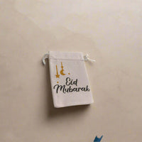 White hessian drawstring bag with 'Eid Mubarak' in black text and gold glitter moon and star decorations, placed on a soft beige marble background with subtle shadows.
