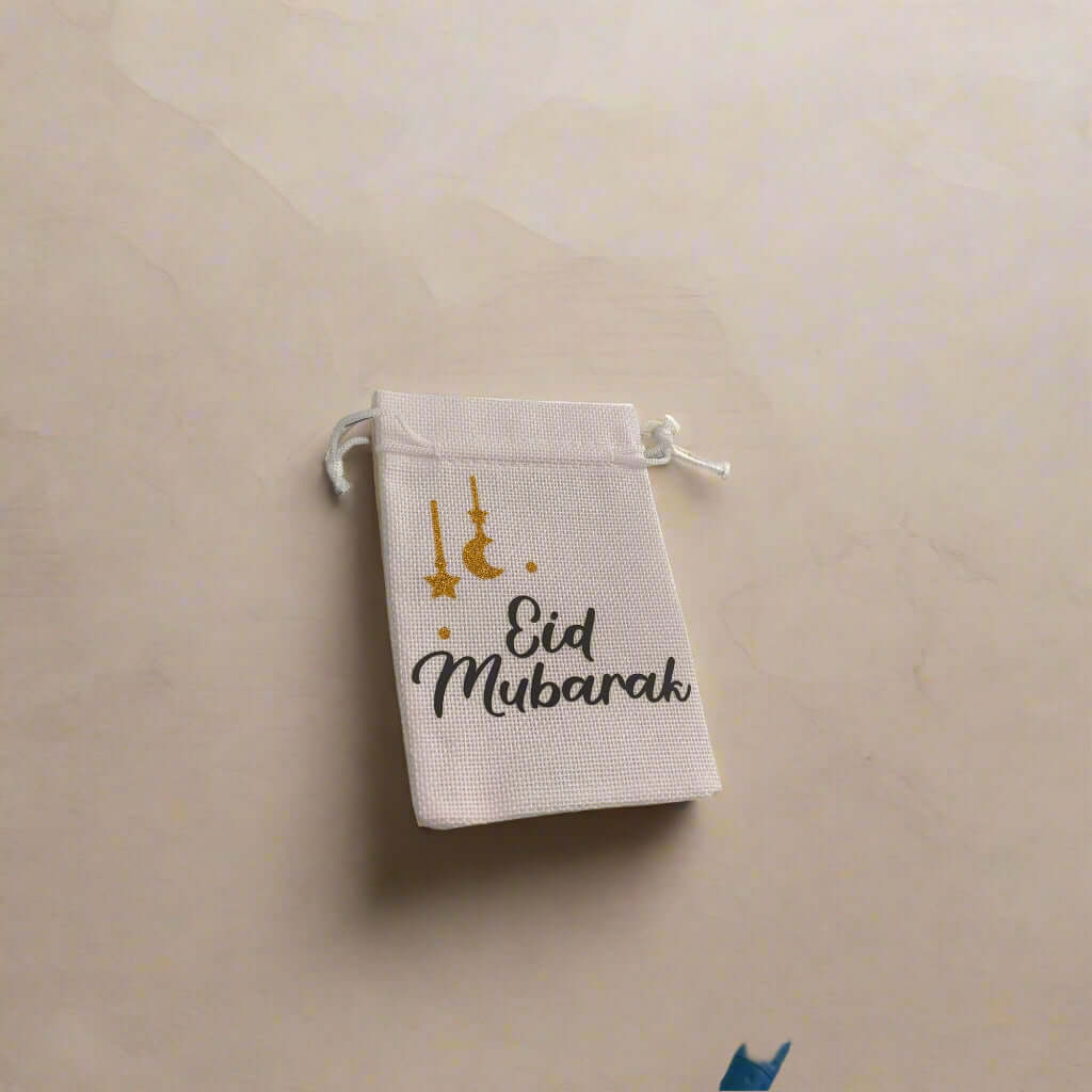 White hessian drawstring bag with 'Eid Mubarak' in black text and gold glitter moon and star decorations, placed on a soft beige marble background with subtle shadows.