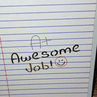 Close-up of encouragement greeting card with A+ Awesome Job message and smiley face on lined paper background, photographed from a straight-on angle.
