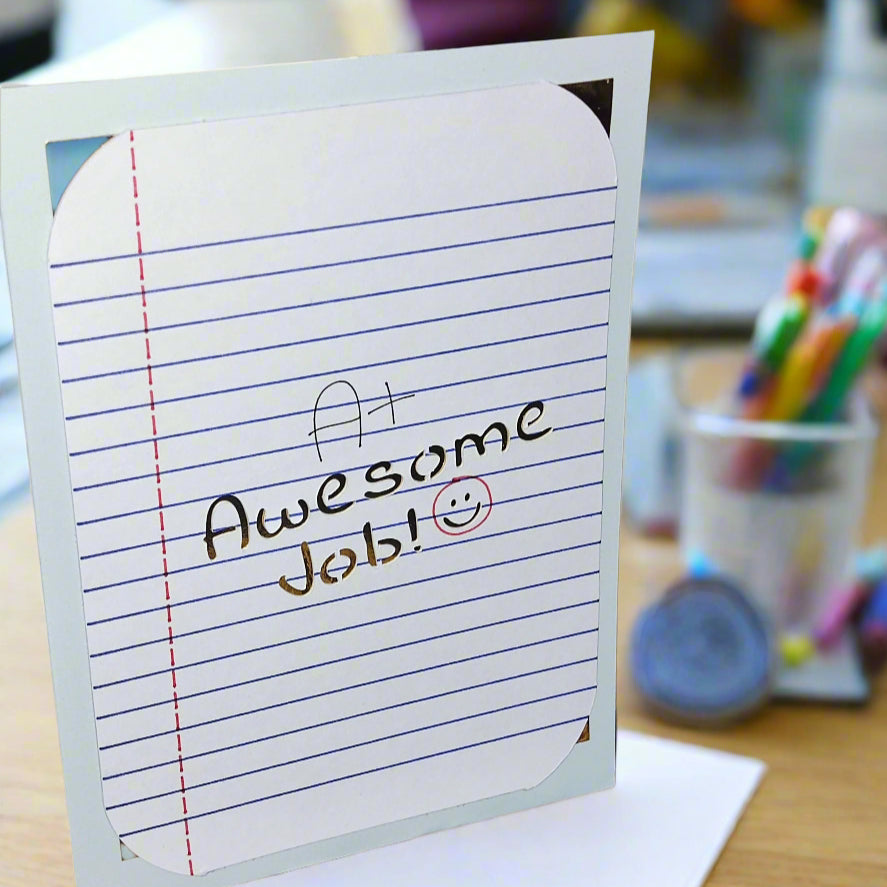 Encouragement greeting card with A+ Awesome Job message and smiley face, displayed upright on a desk with colourful stationery, photographed from a front-side angle.
