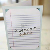 Encouragement greeting card with A+ Awesome Job message and smiley face, standing upright and photographed from a front-facing angle on a blurred neutral background.

