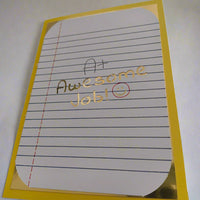 Encouragement greeting card with A+ Awesome Job! message in gold foil on lined paper, mounted on yellow card, photographed from a low side angle.
