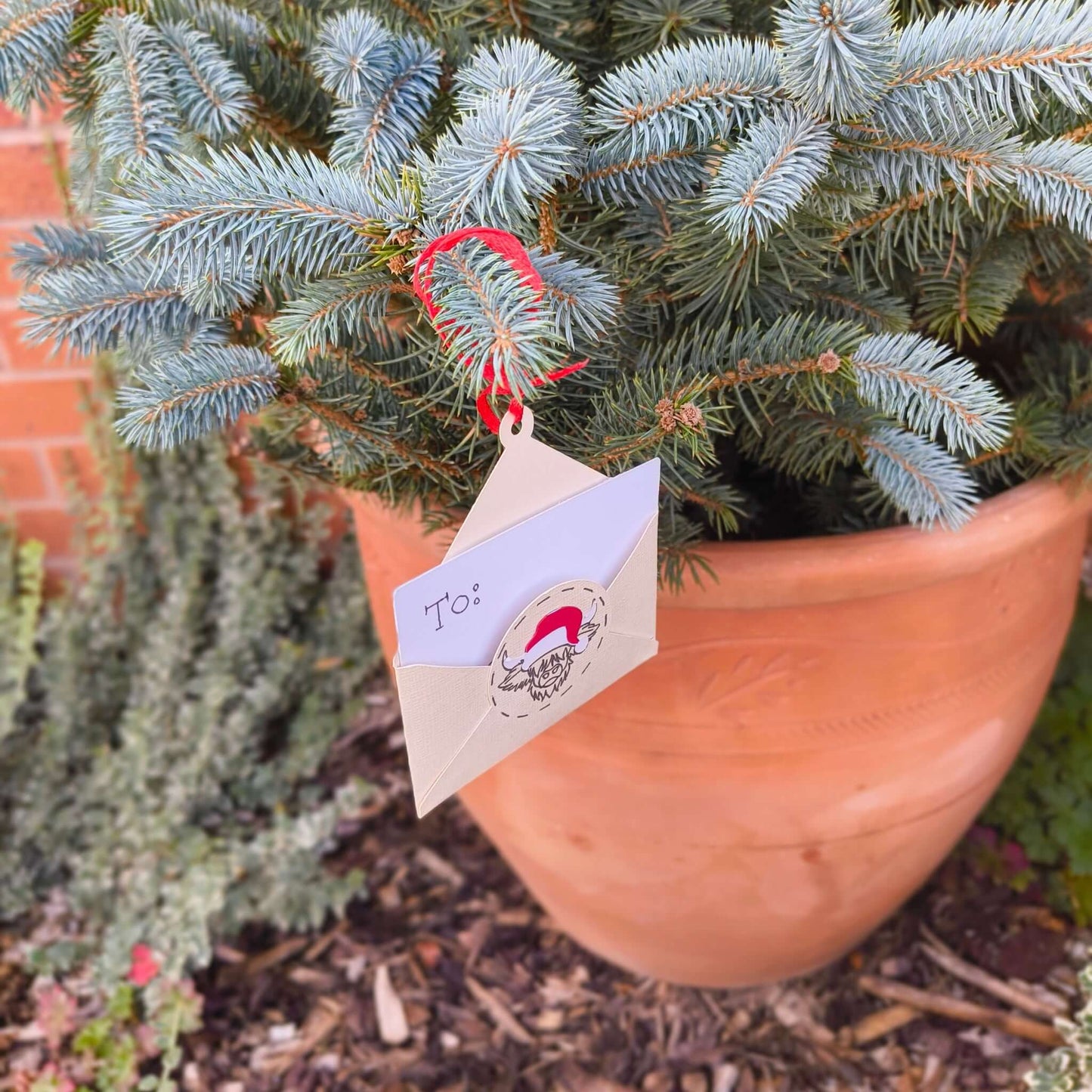 Festive gift card holder envelope ornament with Highland cow Santa design hanging on outdoor tree