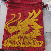 Festive red hessian drawstring bag with gold glitter dragon print and New Year message, flat-lay style.
