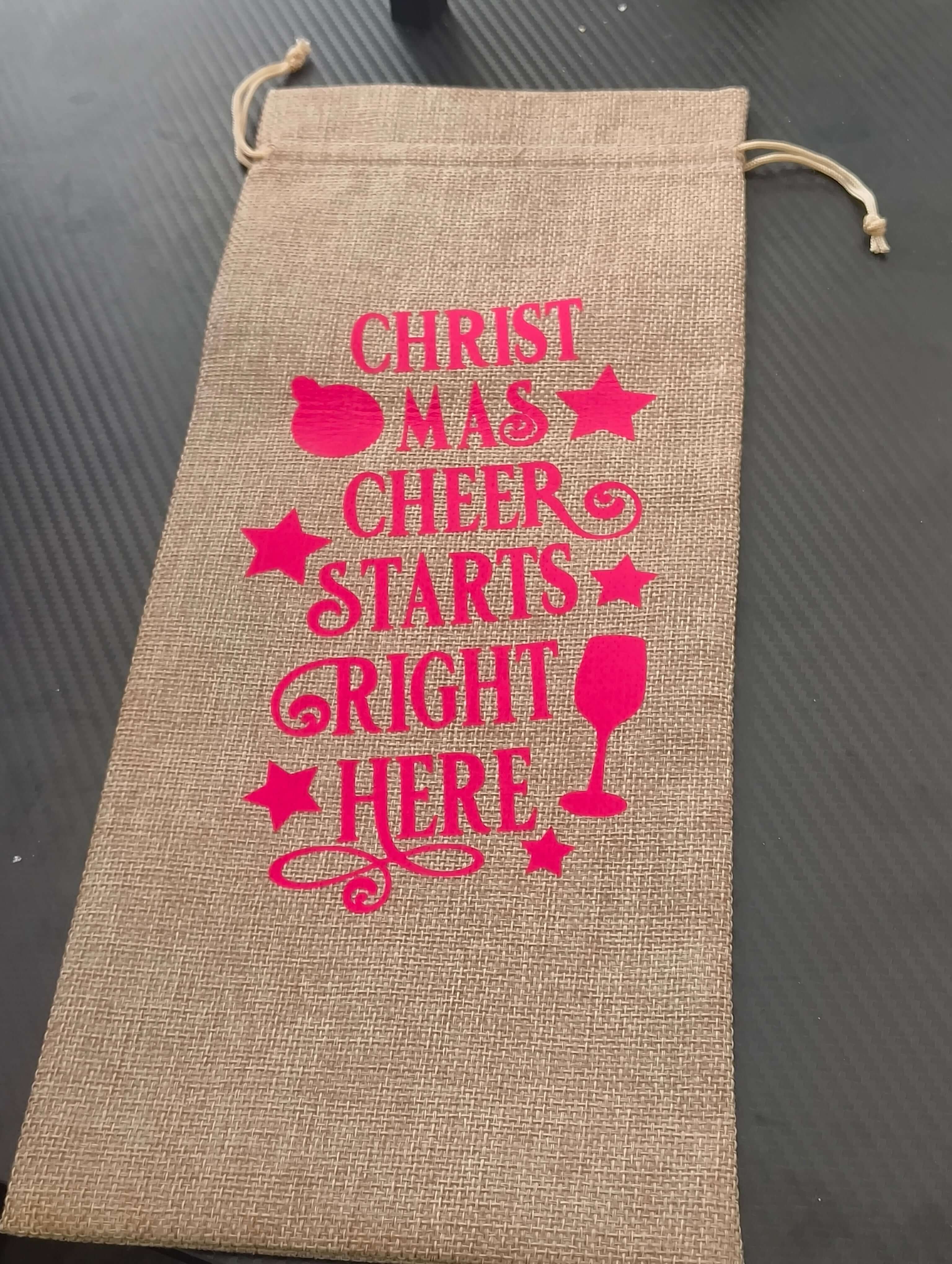 Festive hessian wine bag with pink text saying 'Christmas Cheer Starts Right Here' for eco-friendly gifting.