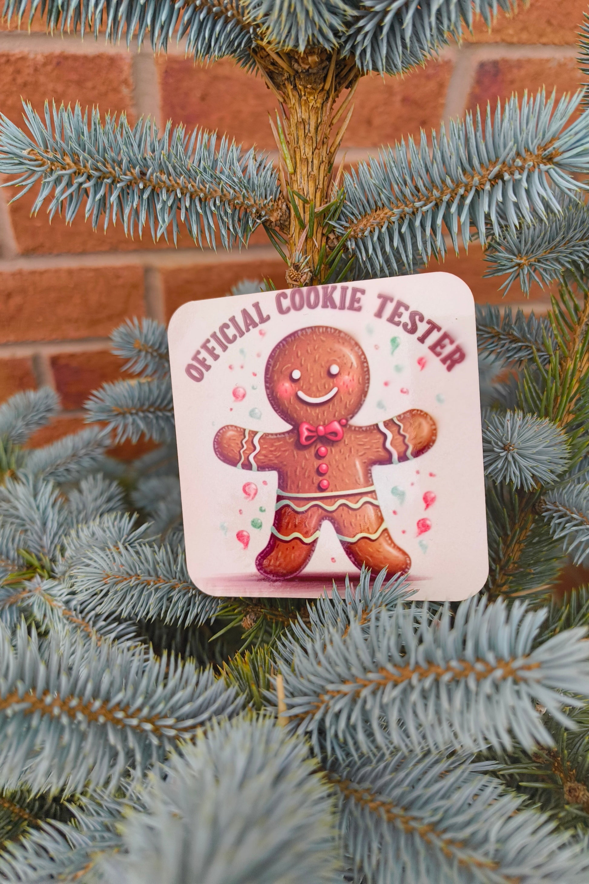 Square coaster with gingerbread man and “Official Cookie Tester” text displayed on a blue spruce tree.