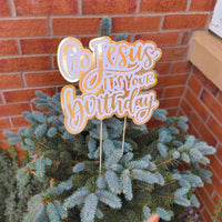Gold and white “Go Jesus It’s Your Birthday” sign styled outdoors in a potted Christmas tree by a brick wall.