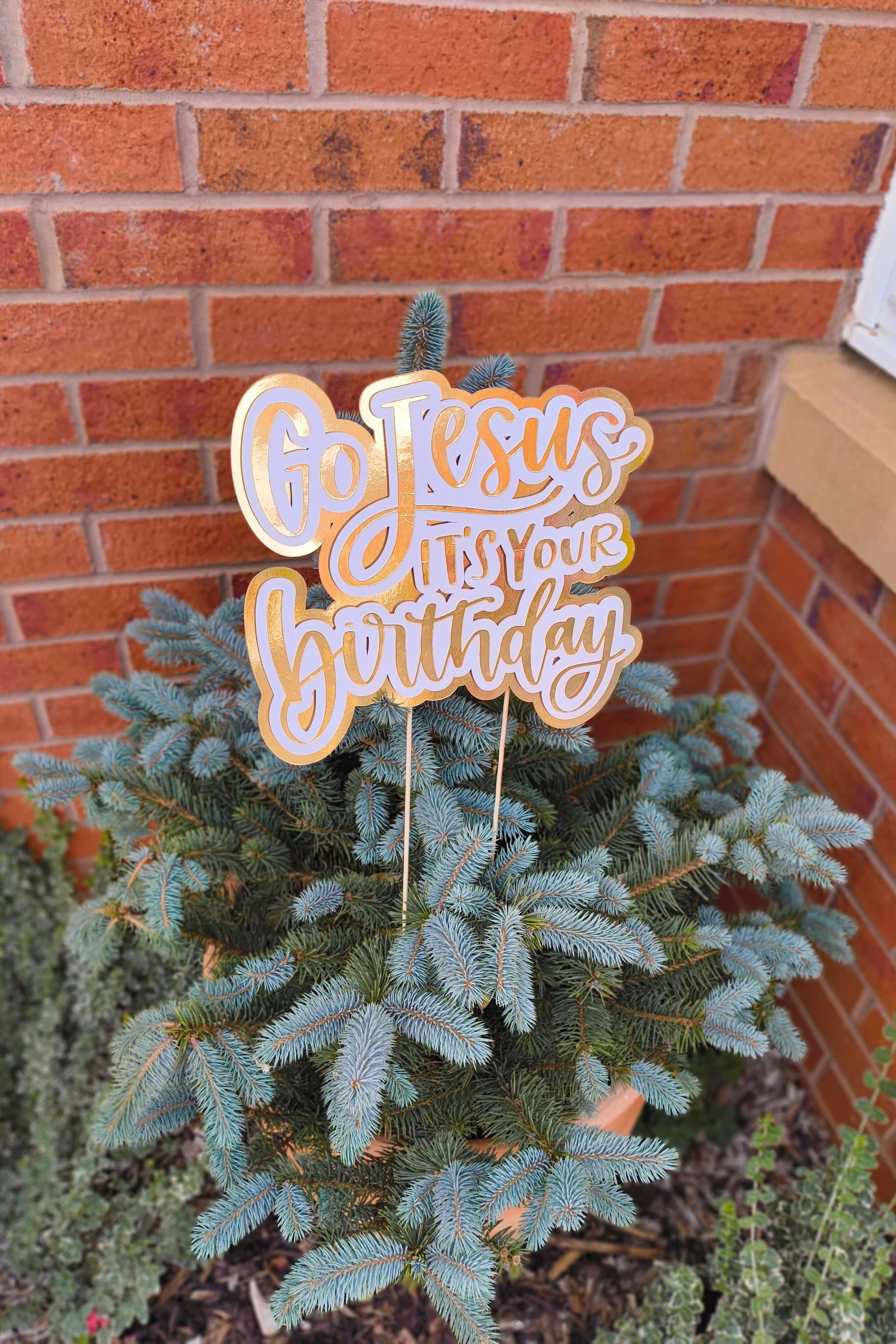 Gold and white “Go Jesus It’s Your Birthday” sign styled outdoors in a potted Christmas tree by a brick wall.
