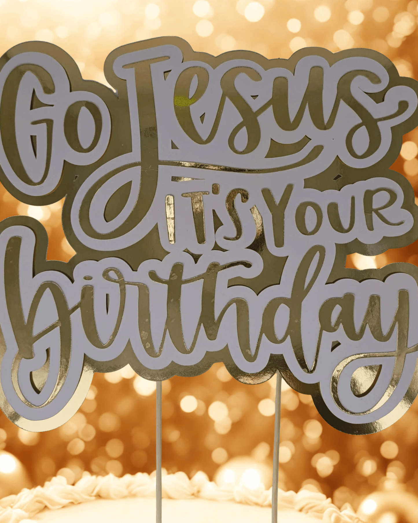 Gold and white “Go Jesus It’s Your Birthday” cake topper on a cake, with golden bokeh background for a festive celebration.