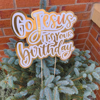 Gold and white “Go Jesus It’s Your Birthday” cake topper displayed on a small blue spruce tree against a brick wall.