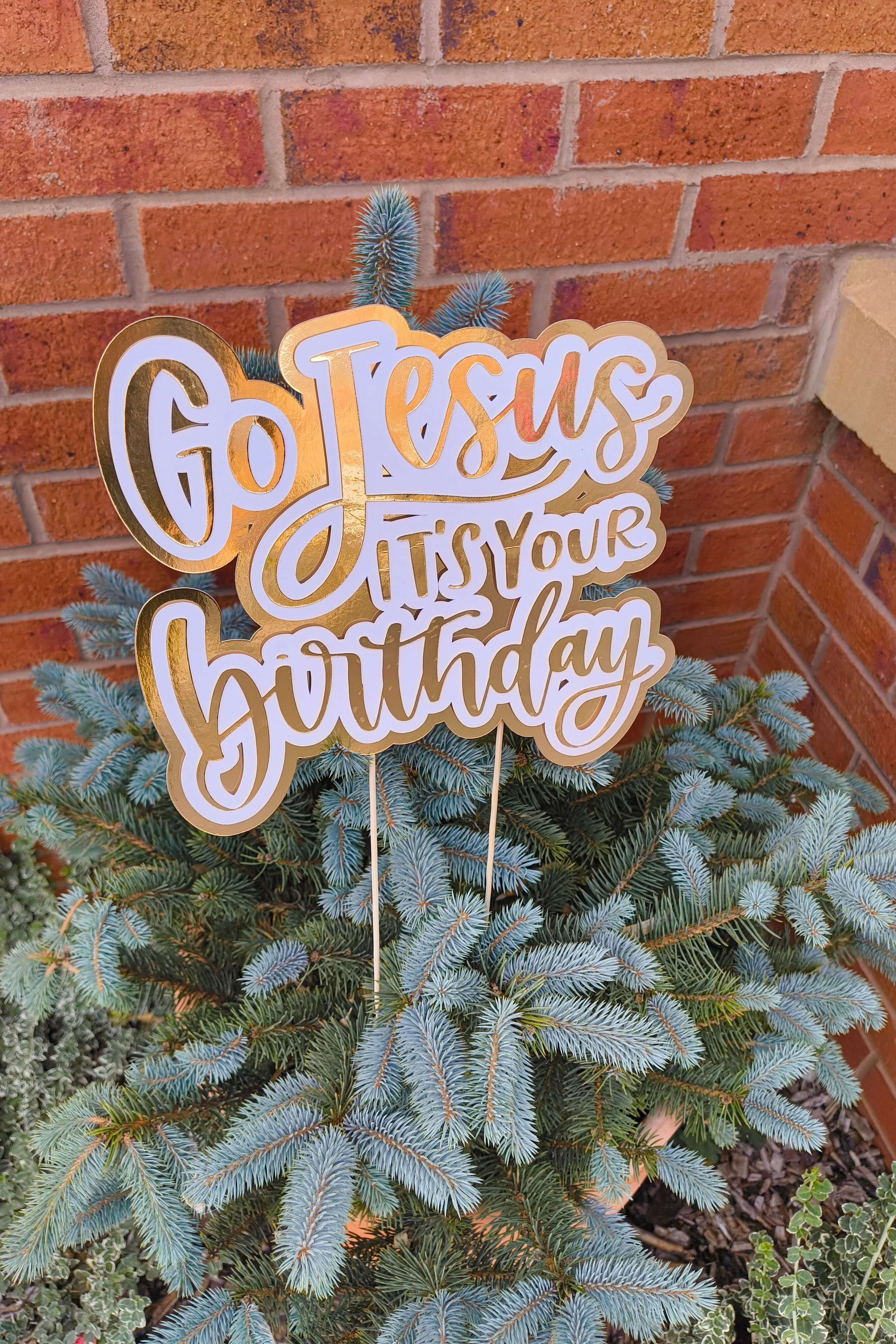 Gold and white “Go Jesus It’s Your Birthday” cake topper displayed on a small blue spruce tree against a brick wall.