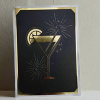 Black and gold greeting card with gold foil cocktail glass and fireworks, mounted on a white backing with gold edges.