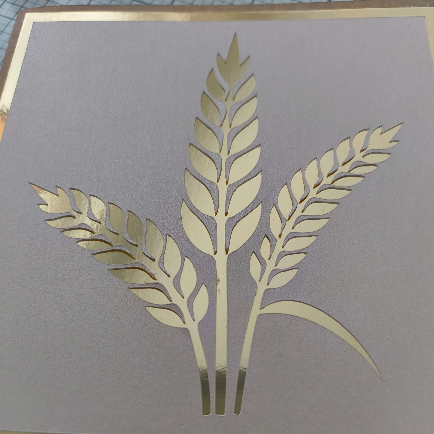 Close-up of a handmade card featuring a metallic gold wheat design on a light beige textured background, with a gold foil border.