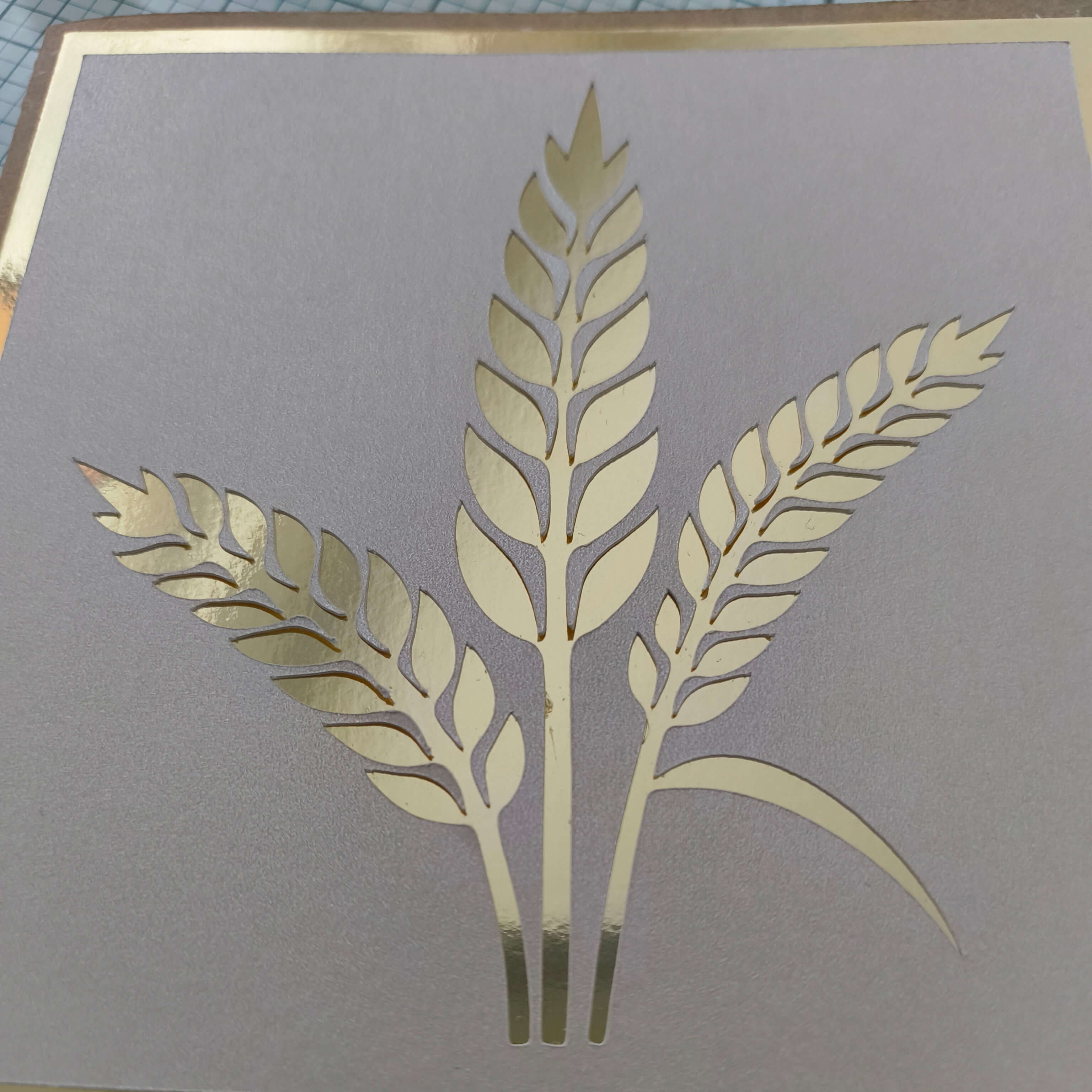 Close-up of a handmade card featuring a metallic gold wheat design on a light beige textured background, with a gold foil border.