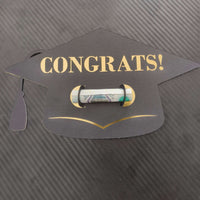 Black graduation cap-shaped money holder with “CONGRATS!” in gold text, holding a rolled banknote, placed on a textured dark background.

