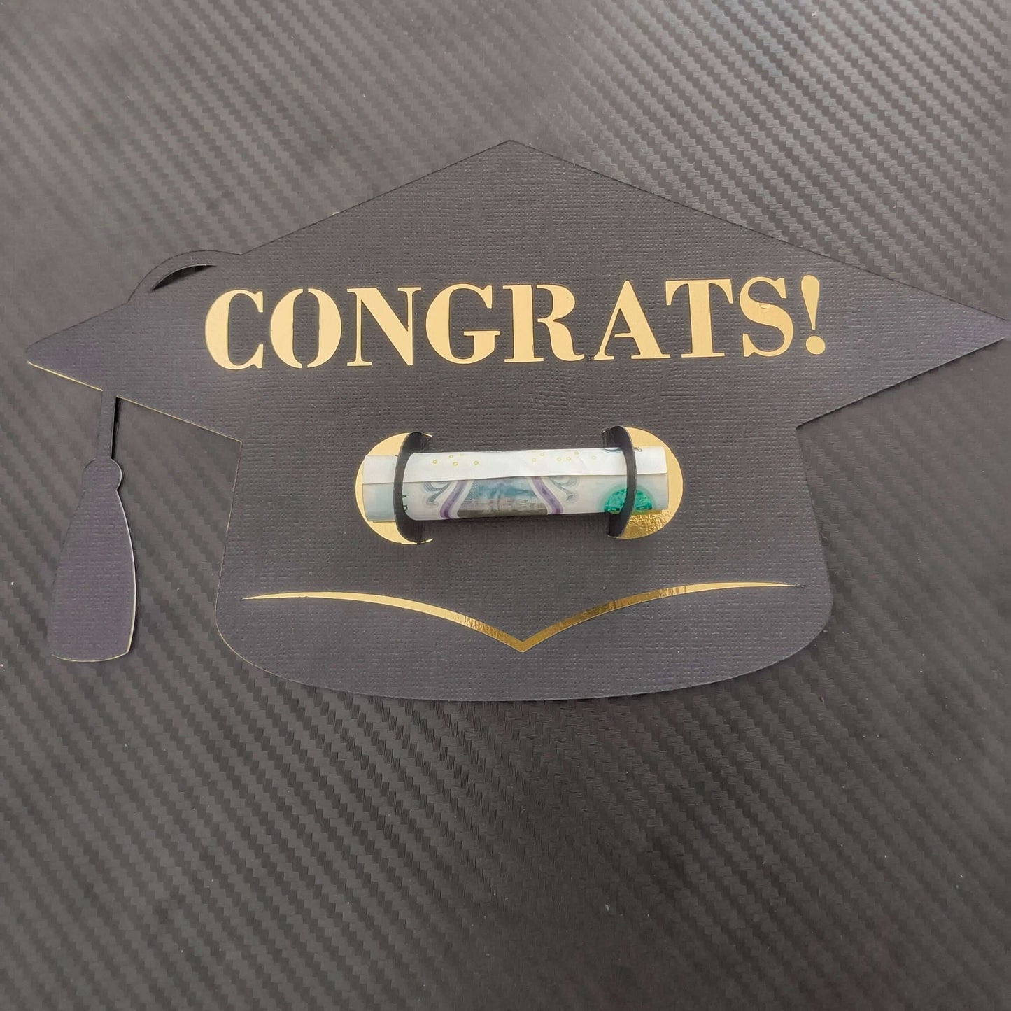 Black graduation cap-shaped money holder with “CONGRATS!” in gold text, holding a rolled banknote, placed on a textured dark background.

