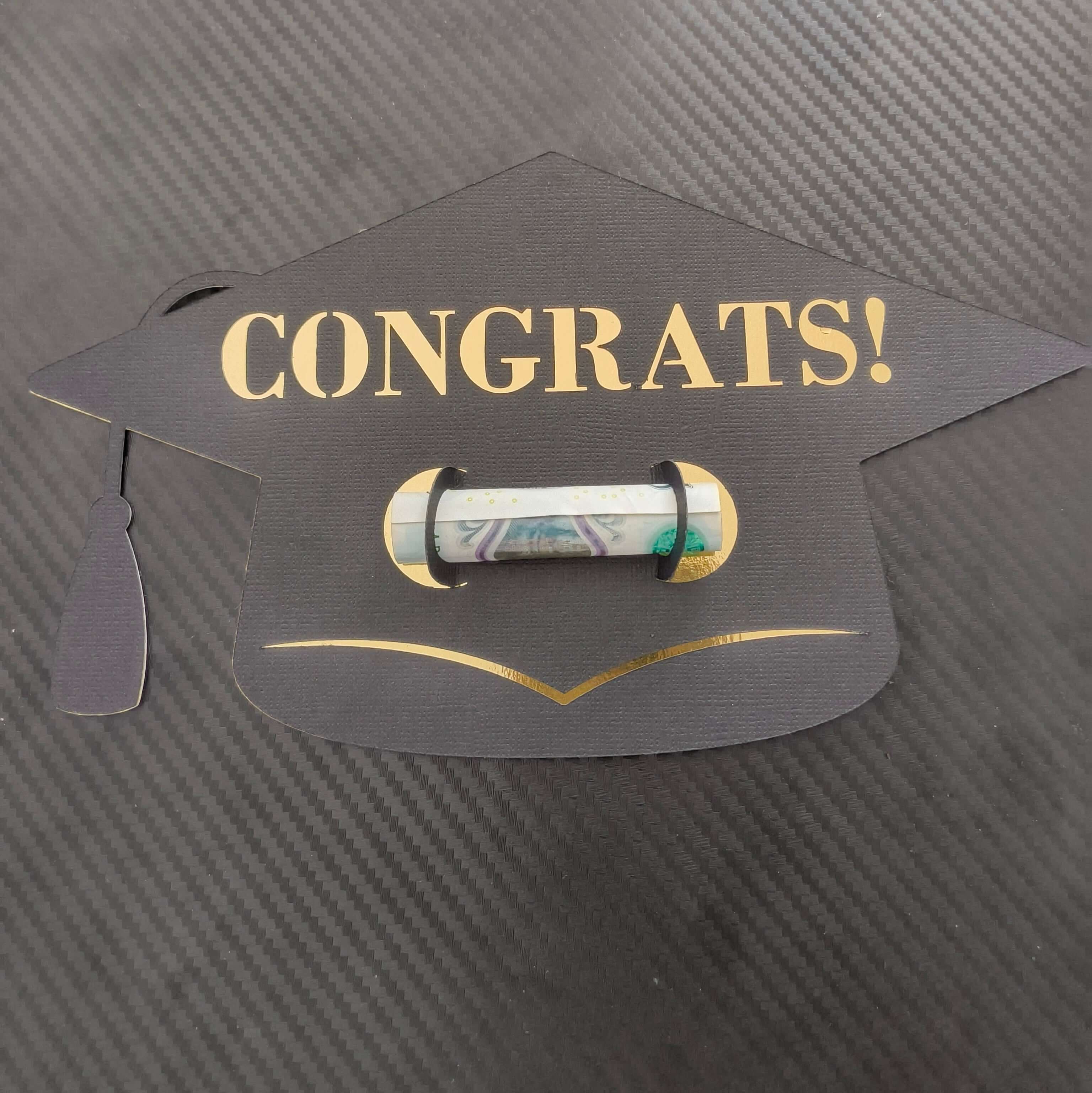 Black graduation cap-shaped money holder with “CONGRATS!” in gold text, holding a rolled banknote, placed on a textured dark background.

