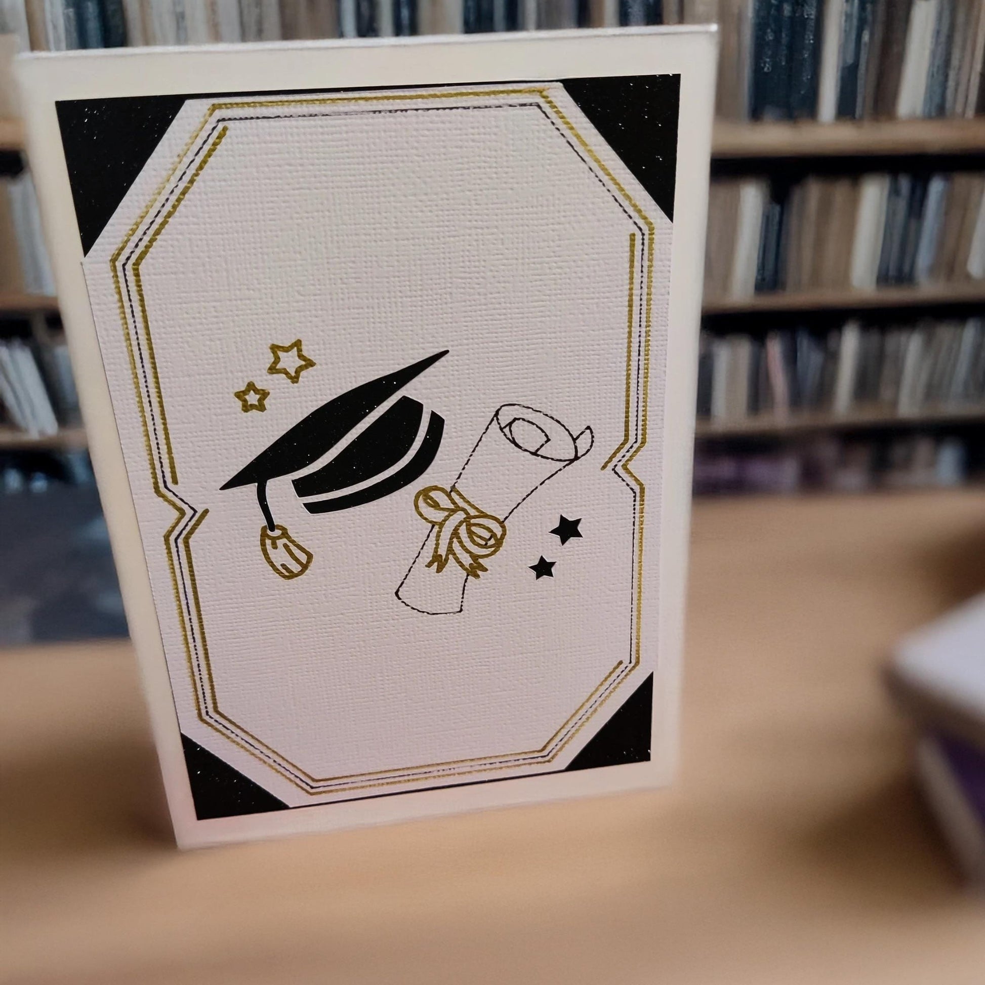 Graduation congratulations card with cap and scroll design in black and gold, displayed standing upright with a bookshelf background.