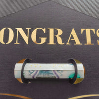 Close-up of black graduation cap-shaped money holder with gold “CONGRATS!” lettering and rolled banknote held securely in the centre slot.

