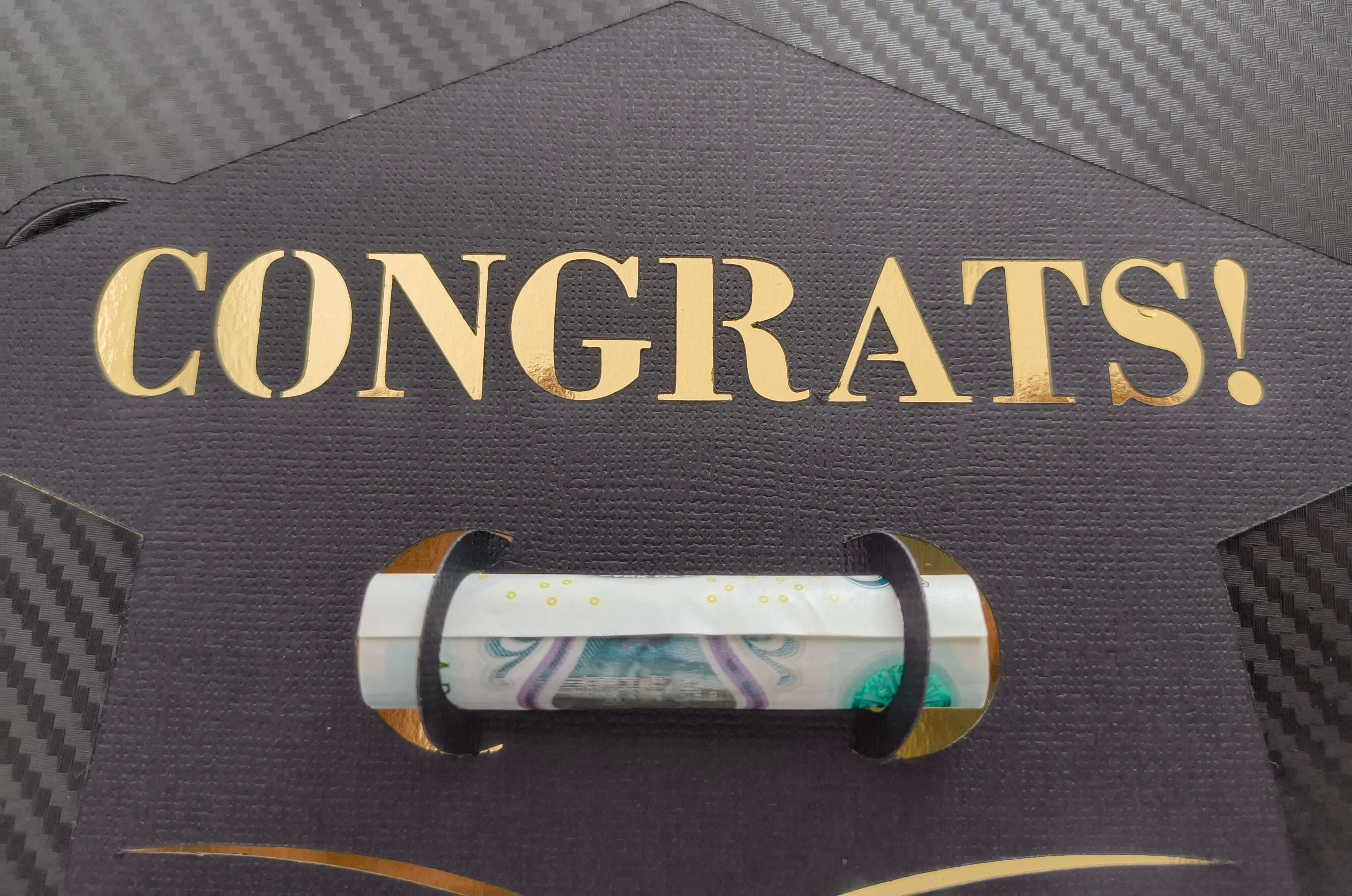 Close-up of black graduation cap-shaped money holder with gold “CONGRATS!” lettering and rolled banknote held securely in the centre slot.

