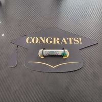 Black graduation cap-shaped money holder with gold “CONGRATS!” text, holding a rolled banknote in the centre, displayed on a dark textured background.