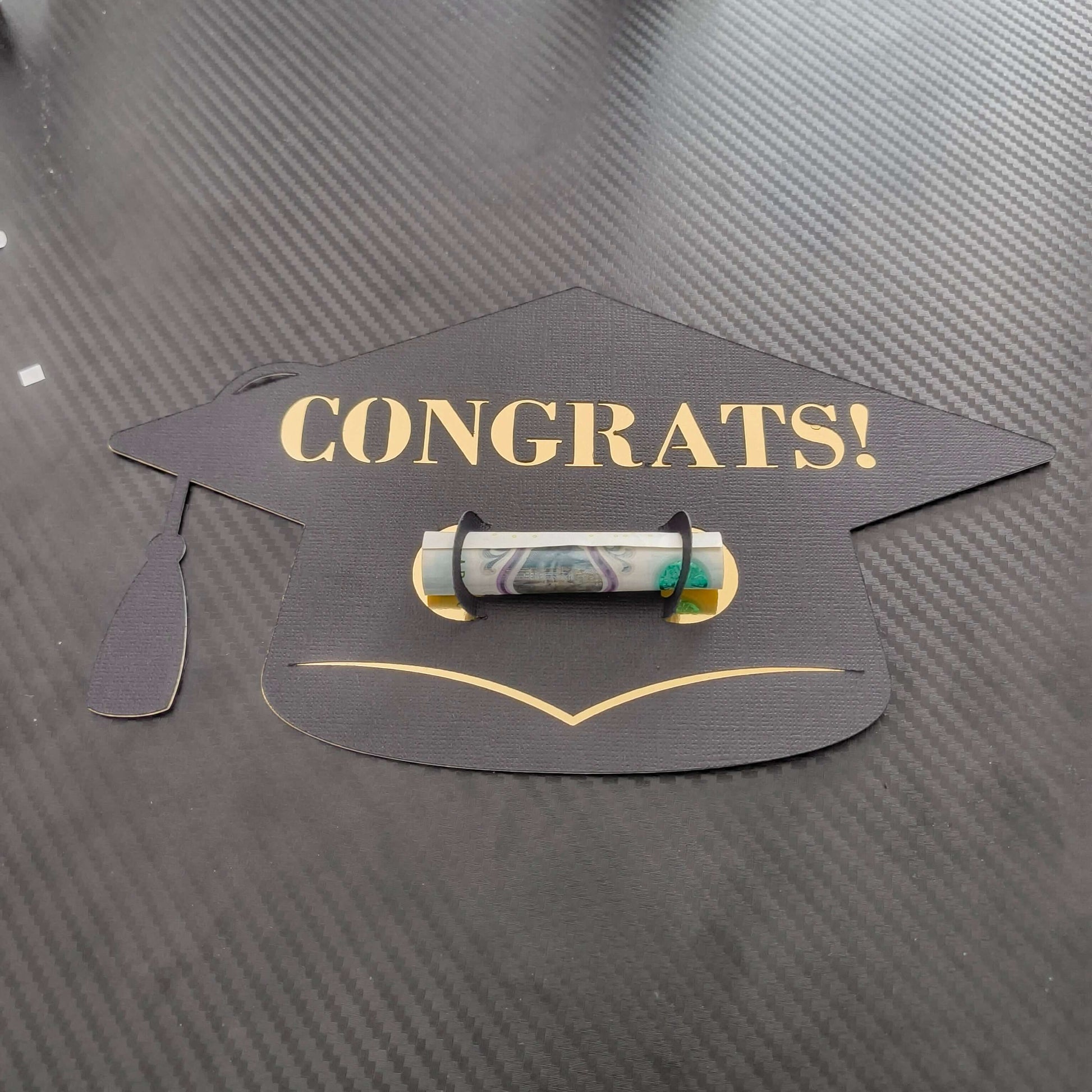 Black graduation cap-shaped money holder with gold “CONGRATS!” text, holding a rolled banknote in the centre, displayed on a dark textured background.