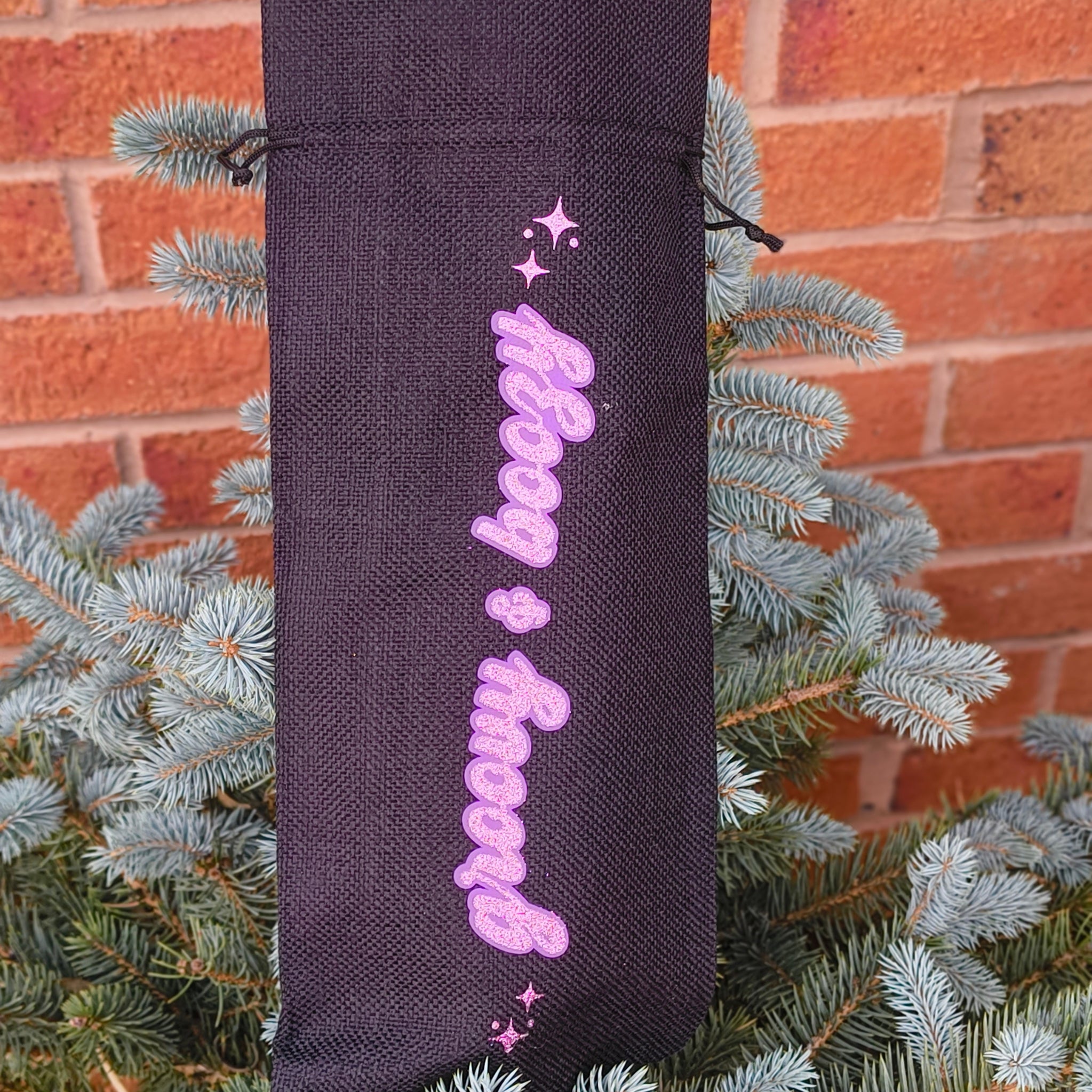 Upright black drawstring wine bag with glitter pink “Groovy & Boozy” lettering, positioned on a pine bush by brick wall.