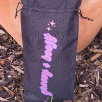 Close-up of black drawstring wine bag with glittery pink “Groovy & Boozy” design and stars, garden background.
