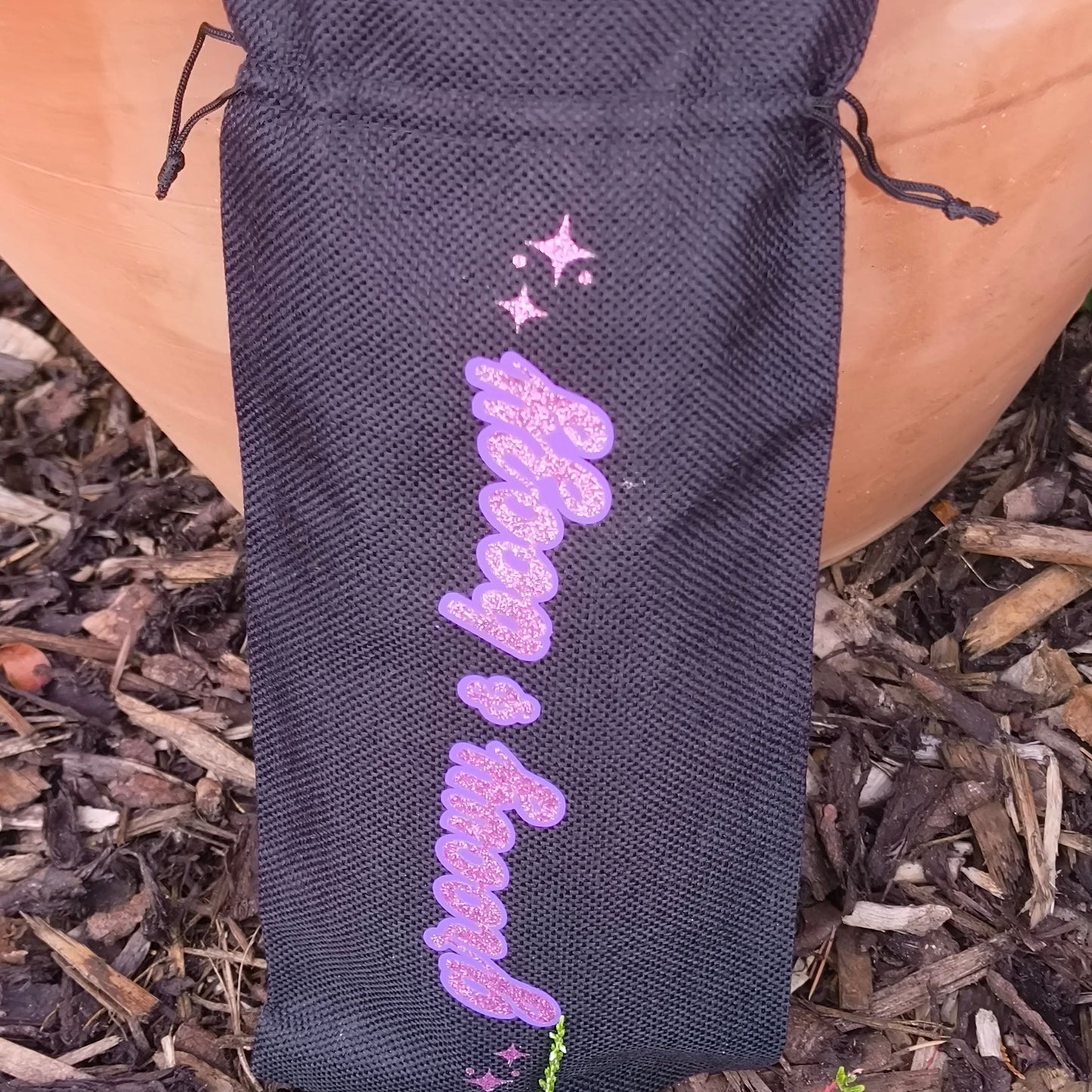 Close-up of black drawstring wine bag with glittery pink “Groovy & Boozy” design and stars, garden background.
