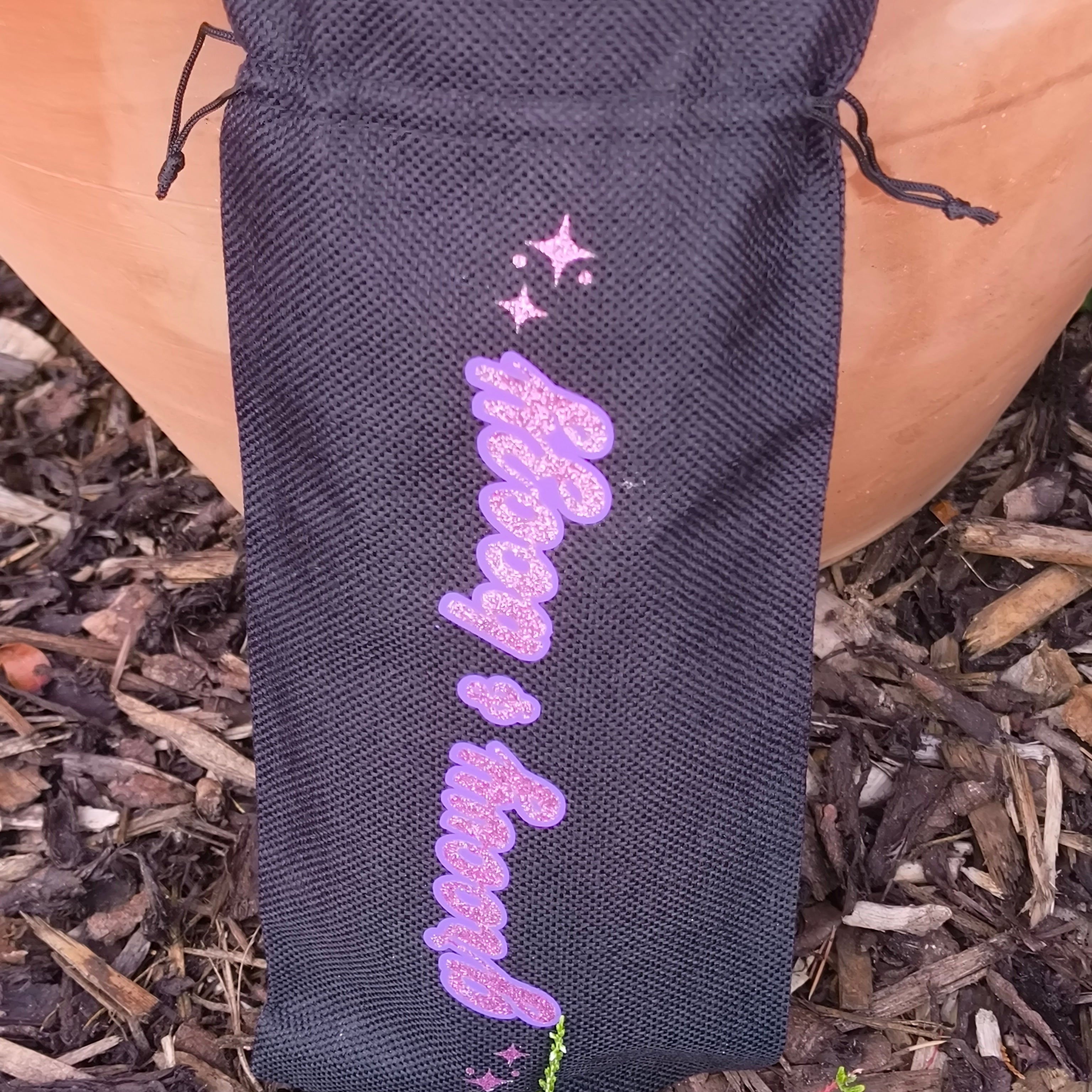 Close-up of black drawstring wine bag with glittery pink “Groovy & Boozy” design and stars, garden background.