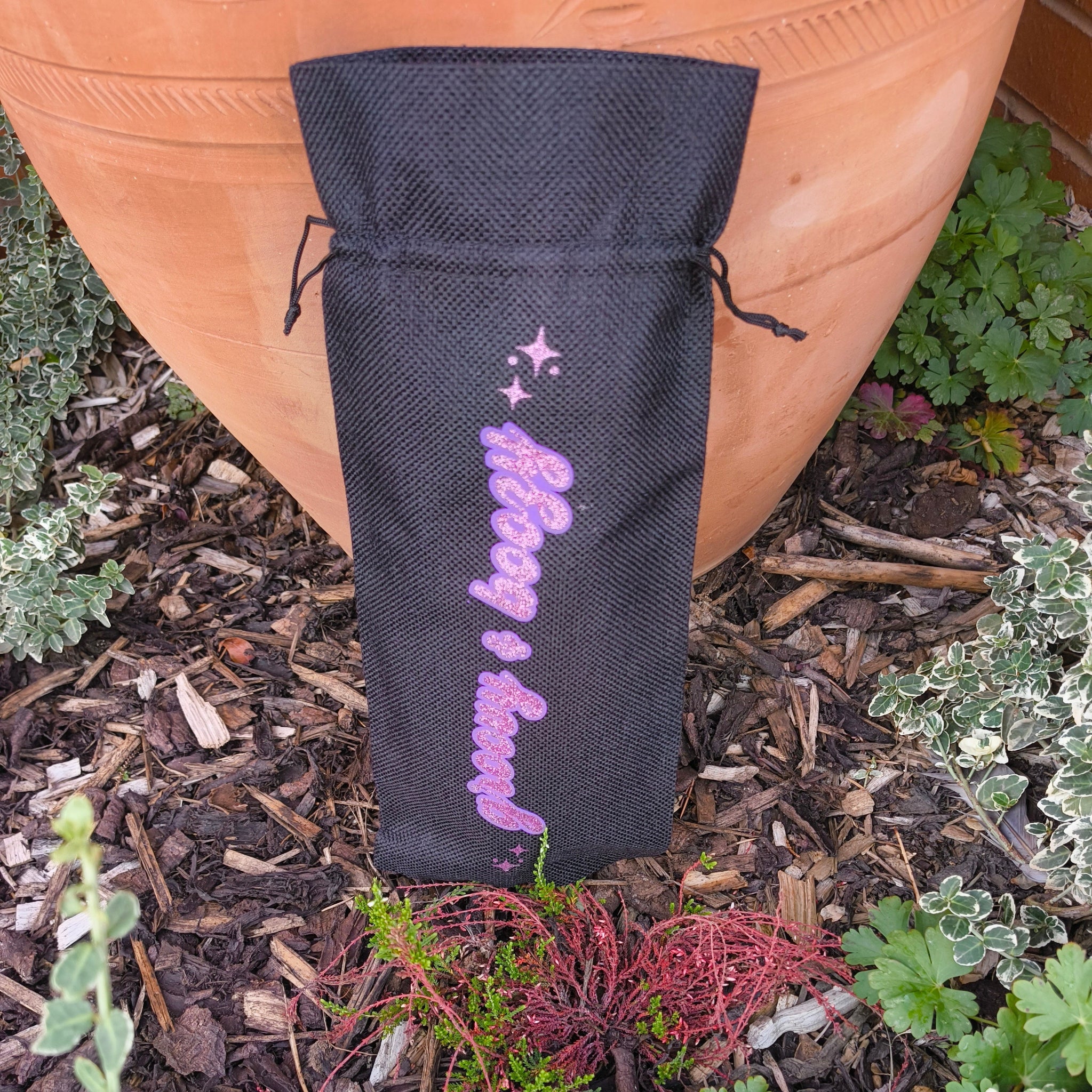 Black wine bag with sparkling pink “Groovy & Boozy” text shown upright outdoors among garden plants.