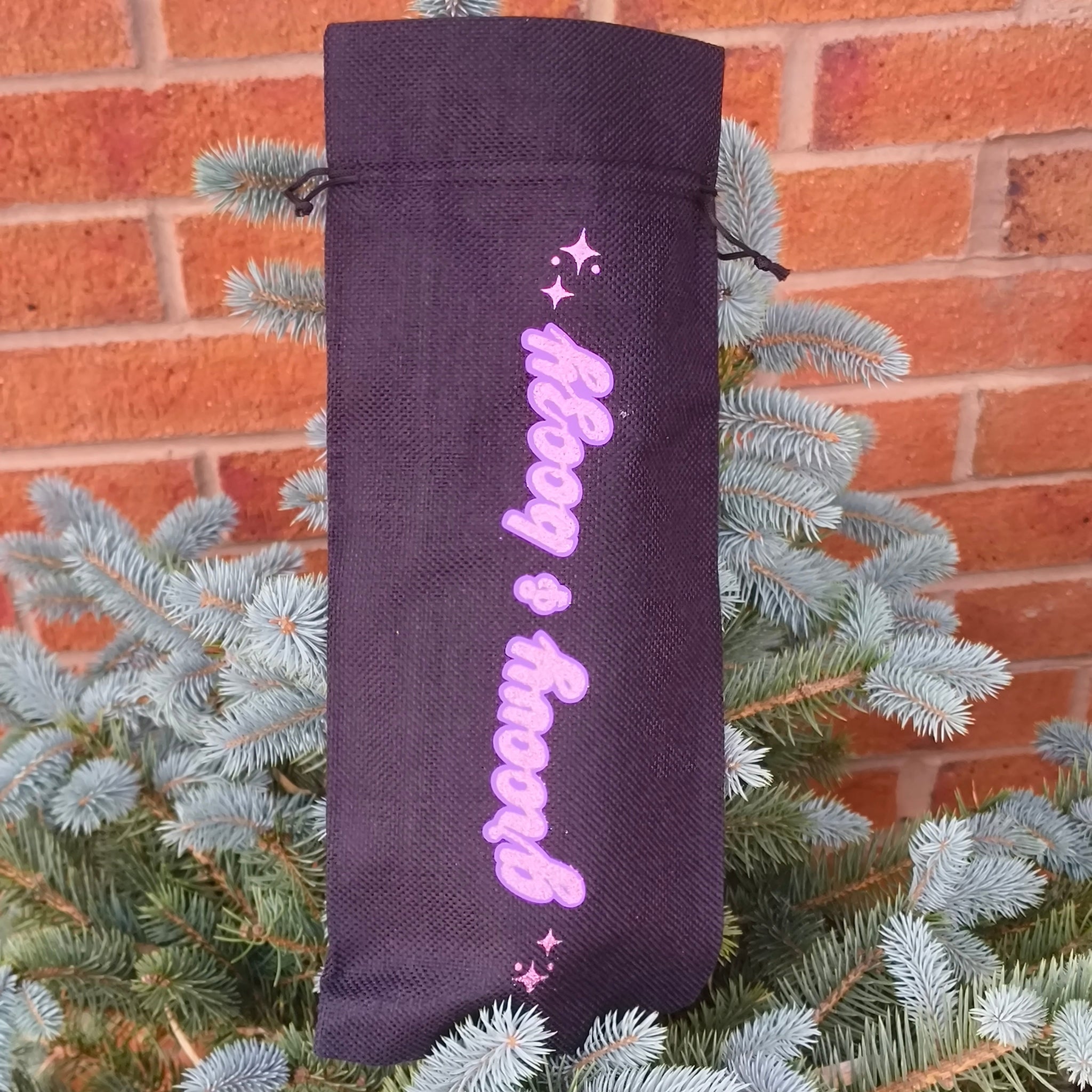 Black wine bag with pink glitter text “Groovy & Boozy” hanging on a small pine tree outdoors near a brick wall.