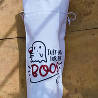 Flat-lay of a Halloween wine bag featuring ghost illustration and glitter red "BOOS" lettering, photographed outdoors on stone tiles.