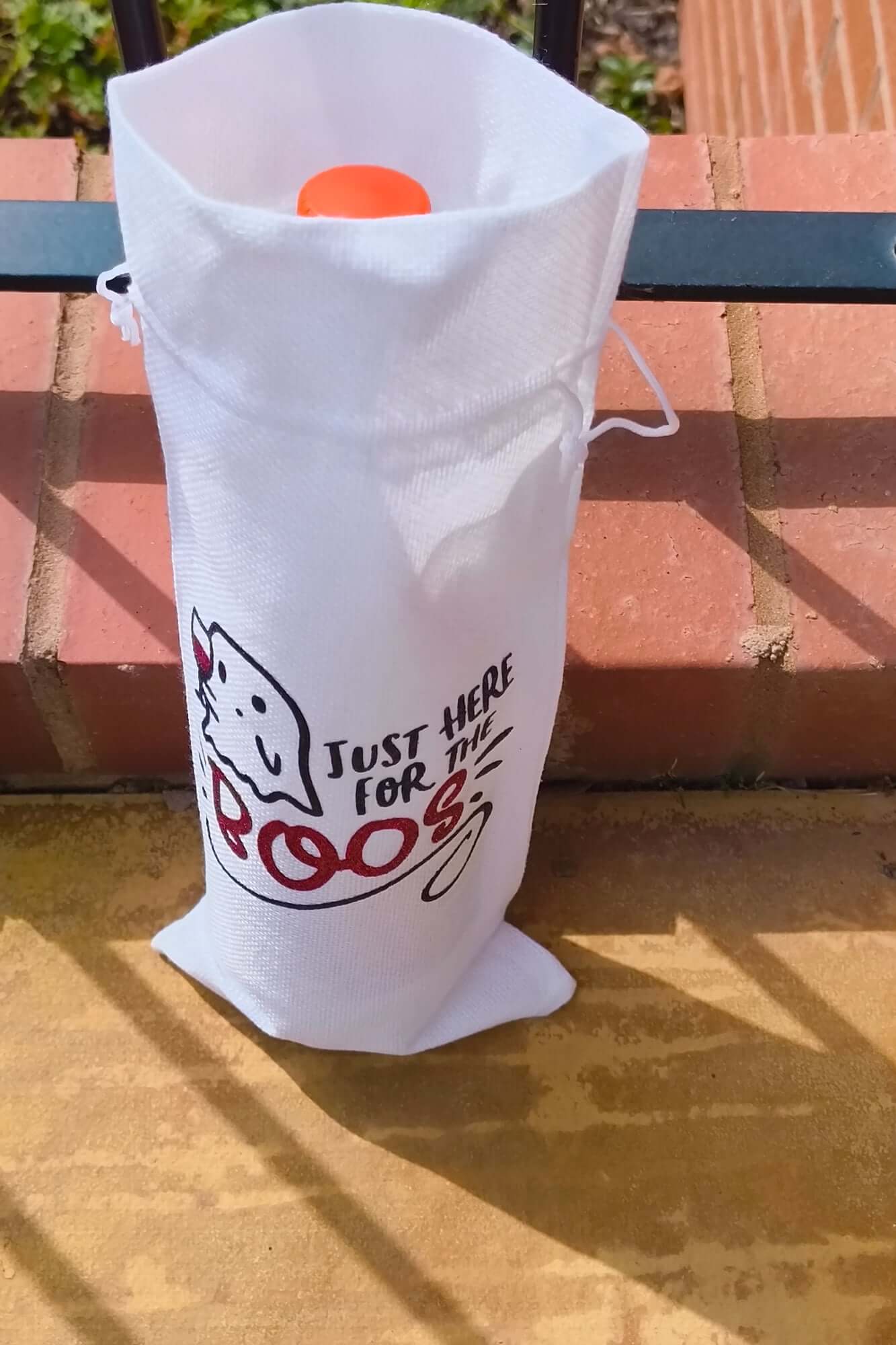 White Halloween wine bag holding a bottle with orange cap, ghost illustration and text "Just Here for the Boos", placed against brick steps.