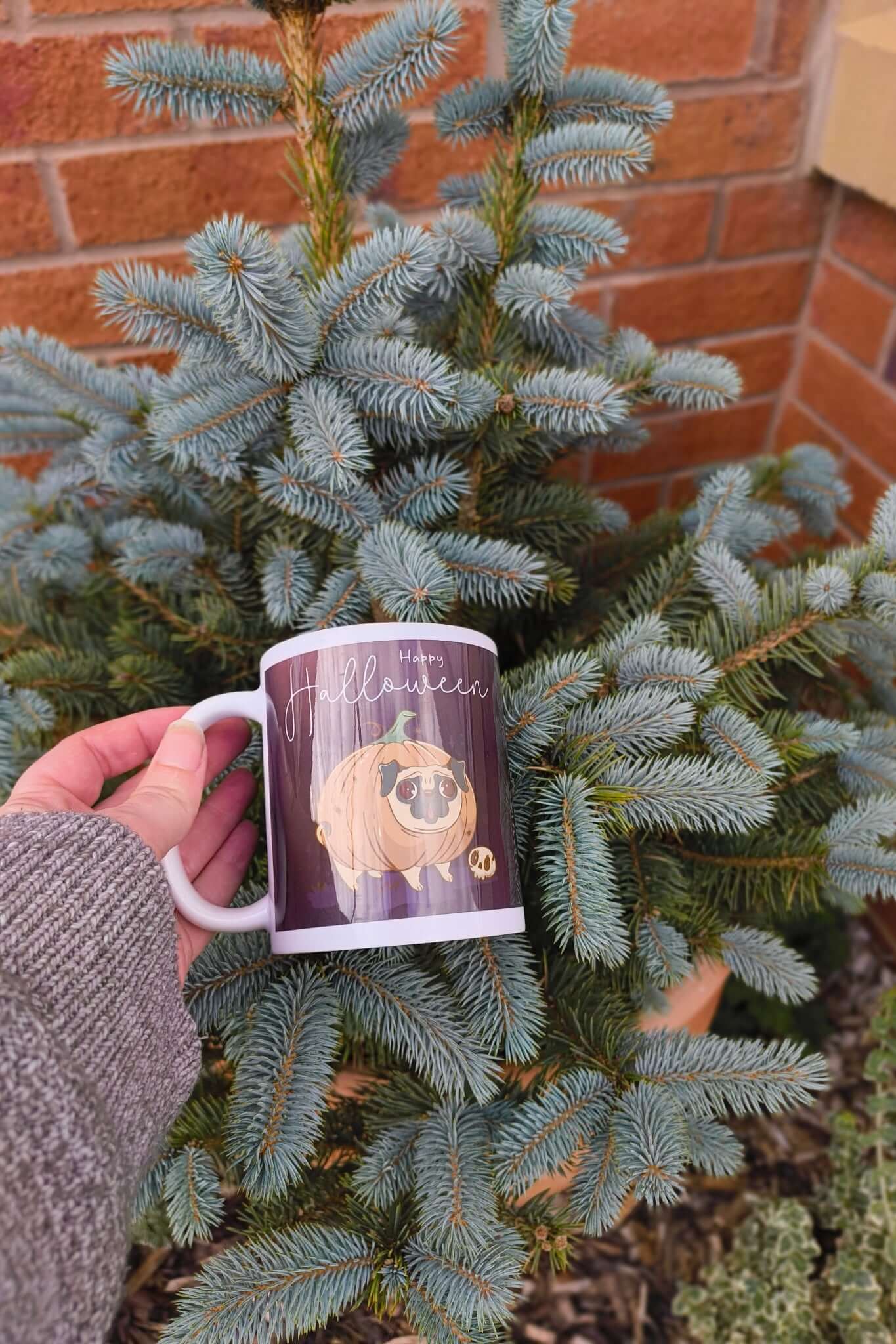 Person holding Happy Halloween mug with pug pumpkin design among fir tree branches, festive autumn outdoor photo.