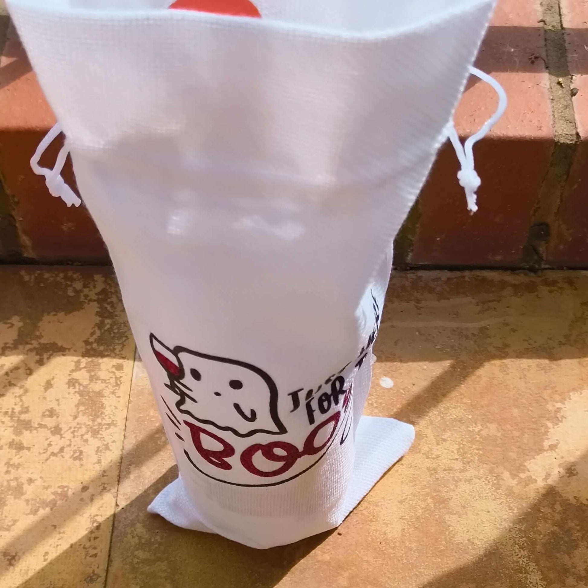 White drawstring wine gift bag with a ghost and "Just Here for the Boos" design, standing upright on outdoor tiles.