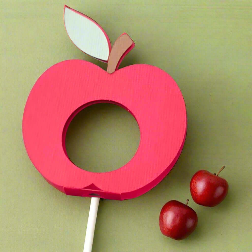 Handmade red apple-shaped lollipop holder with green leaf and brown stem, made from textured cardstock, displayed on a dark carbon-textured background.

