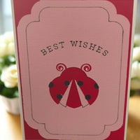 Handmade “Best Wishes” greeting card featuring a red ladybird on textured pink cardstock, displayed upright with soft floral background—ideal for birthdays, congratulations, or general messages.