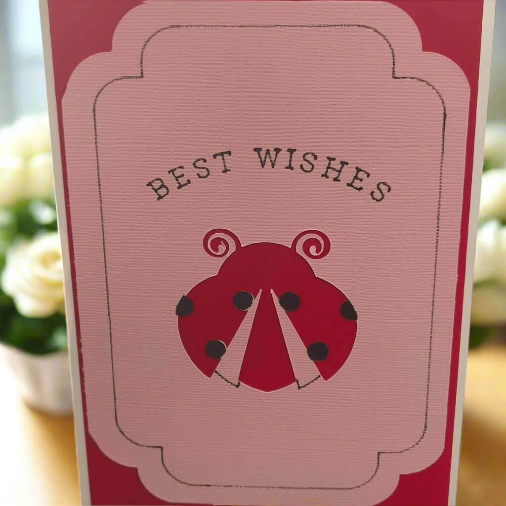 Handmade “Best Wishes” greeting card featuring a red ladybird on textured pink cardstock, displayed upright with soft floral background—ideal for birthdays, congratulations, or general messages.