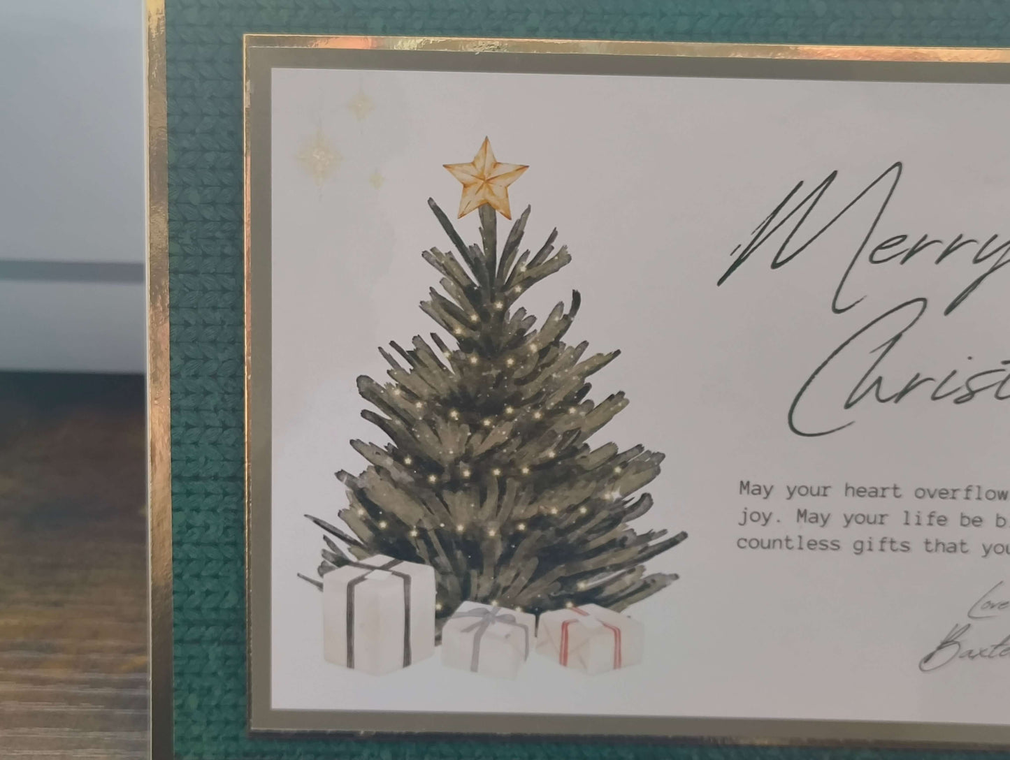 Close-up of the left side of a handmade Christmas card featuring a decorated tree with warm white lights and a gold star topper. The base of the tree is surrounded by wrapped presents in white paper with red and black ribbons. The card is bordered in deep green textured cardstock and gold foil trim.