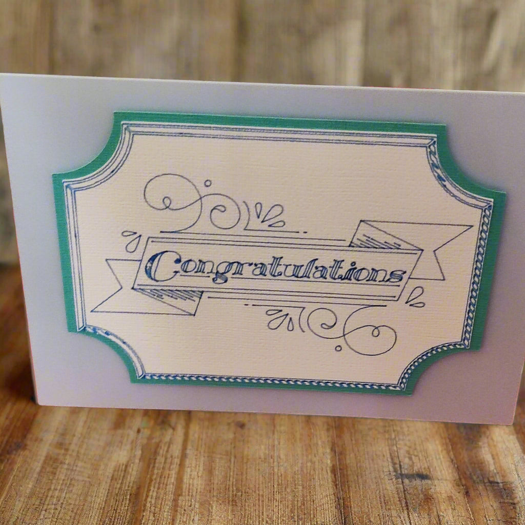 Handmade congratulations card with a vintage-style banner and teal border. Textured ivory cardstock adds a tactile finish. Standard greeting card size, displayed on a rustic wooden table.
