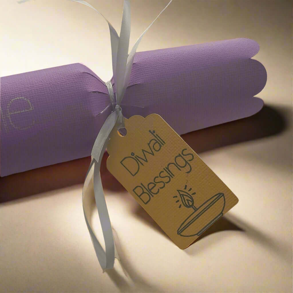 Handmade purple Diwali cracker with white ribbon and golden tag reading 'Diwali Blessings' featuring a diya illustration, displayed on a softly lit background.