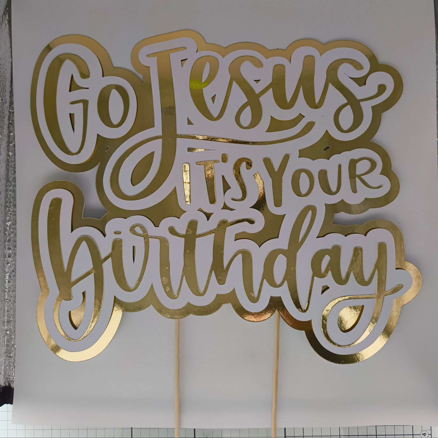 Handmade cake topper with gold lettering reading “Go Jesus It’s Your Birthday,” mounted on wooden sticks for decoration.
